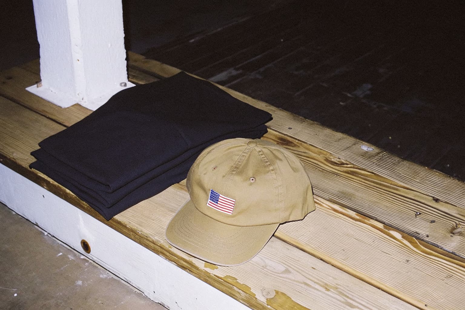 Folded black t-shirt and a military green ballcap with an American flag patch stacked on a dark wood plank