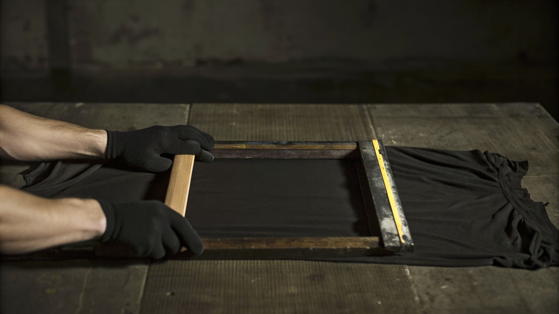 Gloved hands pulling a screen printing squeegee across a mesh screen onto a black t-shirt in the shop