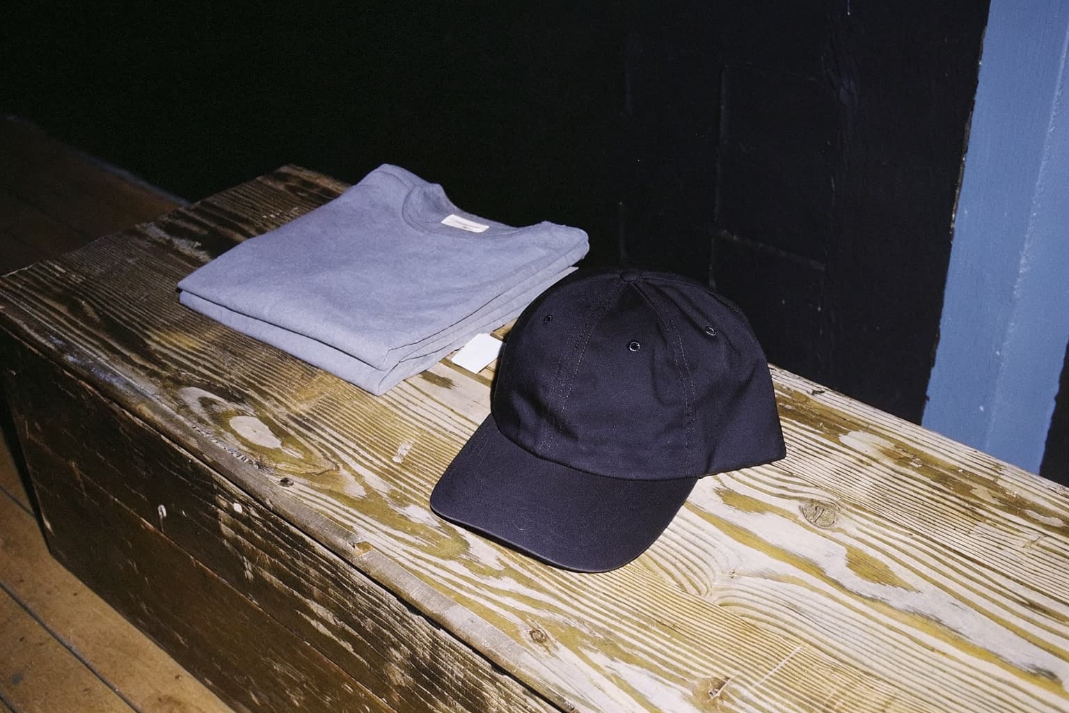 A folded black graphic t-shirt and a black embroidered trucker hat on a dark wood plank surface